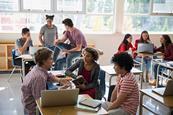 Photo of a classroom with some students sitting at their desks and others standing.